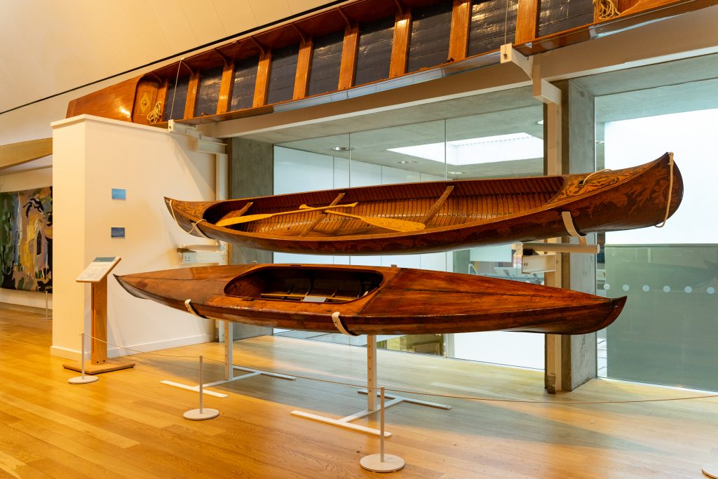 Three wooden boats on display in a gallery. The middle one is highly ornately decorated with inlaid wood and all are varnished.