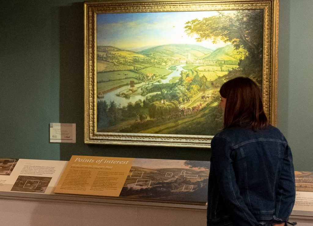 A woman with dark hair is looking at a large oil painting of Henley in a gilt frame, in front of which is an information panel indicating familiar features in the town today.