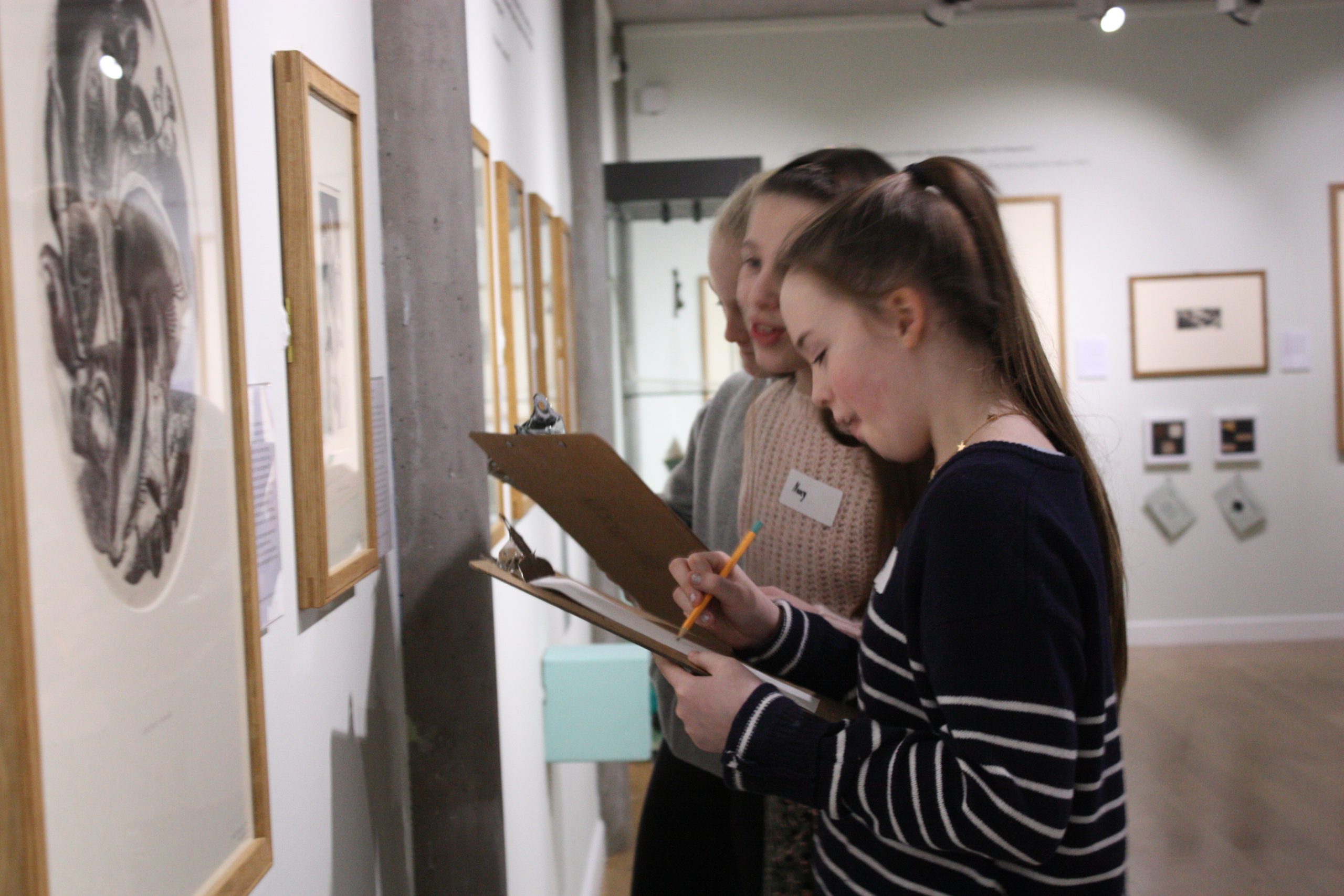 Three teenage girls are drawing from artworks on the gallery wall, using clipboards.