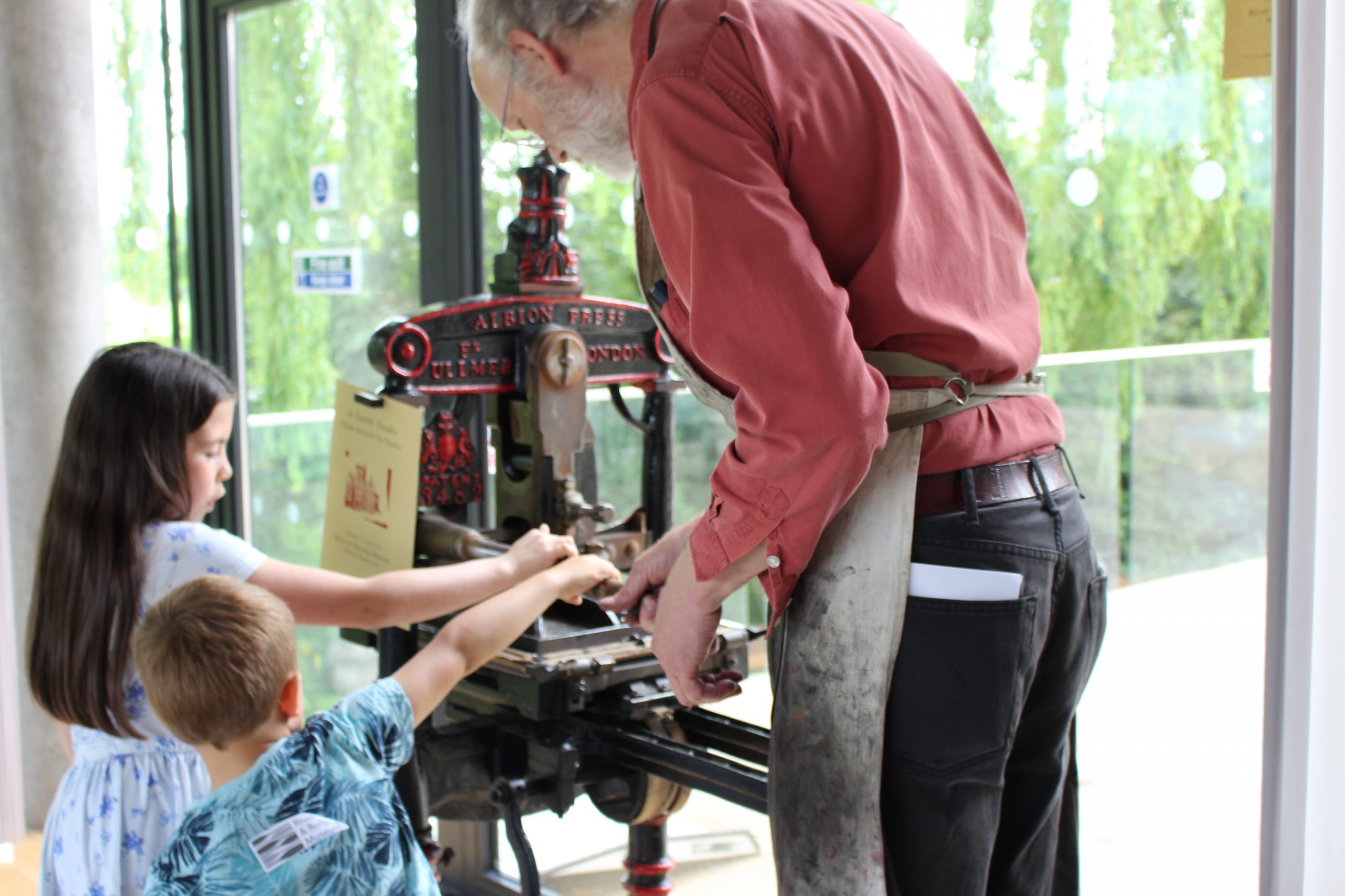 A young girl and boy together turn the handle to operate an old-fashioned printing press. They are helped by an older man wearing an ink-stained apron