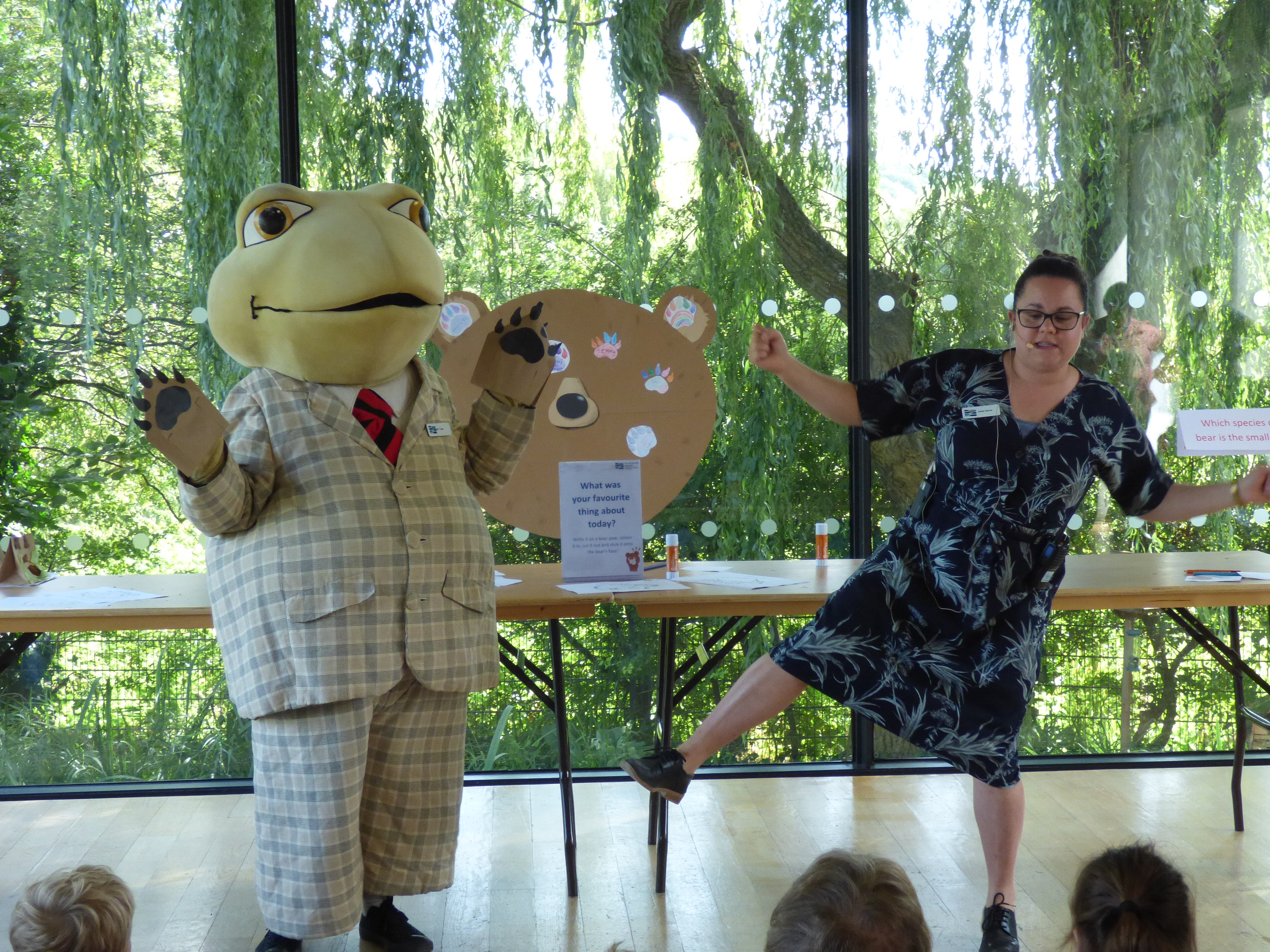 A woman is doing a dance with a Mr Toad character in front of an audience of children. The backdrop is leafy willow branches in the sunlight