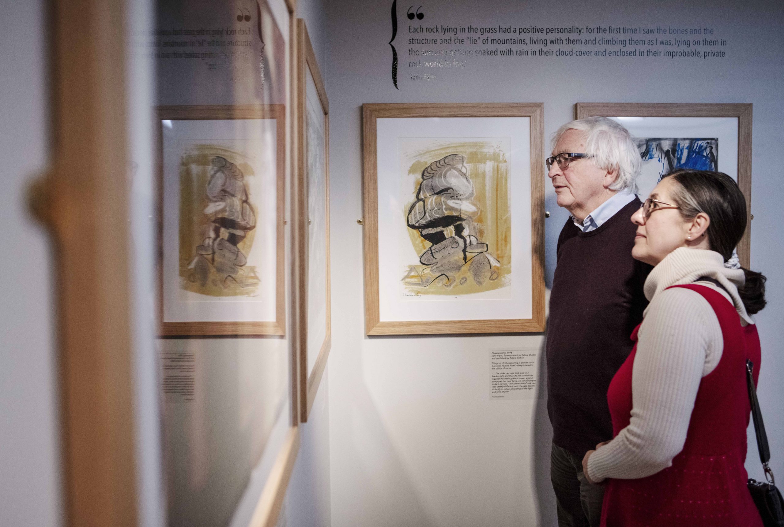 A man and a woman are looking at artworks on the wall in the Piper Gallery. A yellow and grey artwork beside them is reflected in the glass of other artworks.