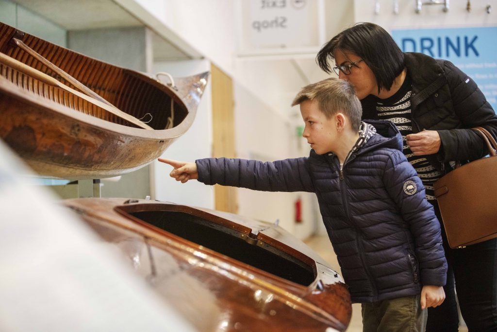 A boy and his mother lean over to look intently at a decorated wooden boat on display in the Museum. The boy is pointing at it.