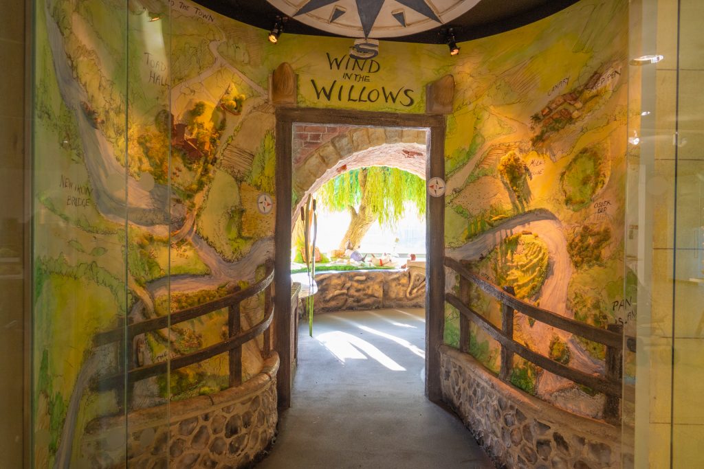 The entrance to the Wind in the Willows experience, with the doorway surrounded by a low wall and fence, and surrounded by a 3D map. Through the door, light streams through around a model willow tree.