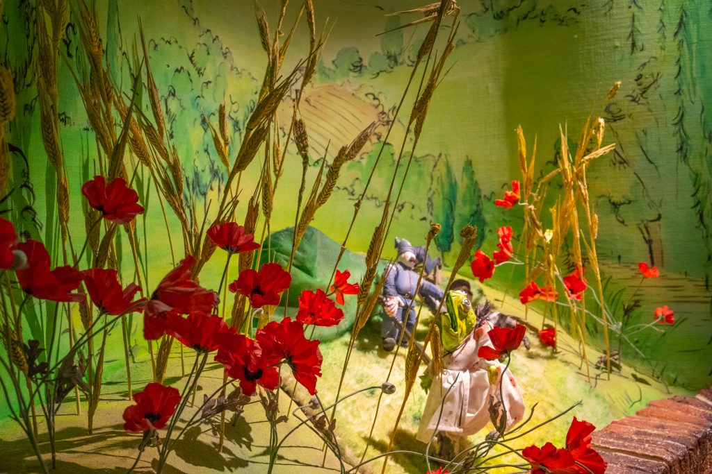 A scene from the Wind in the Willows experience with models of Mr Toad wearing a washer woman's dress, and two police men, seen through models of poppies and wheat