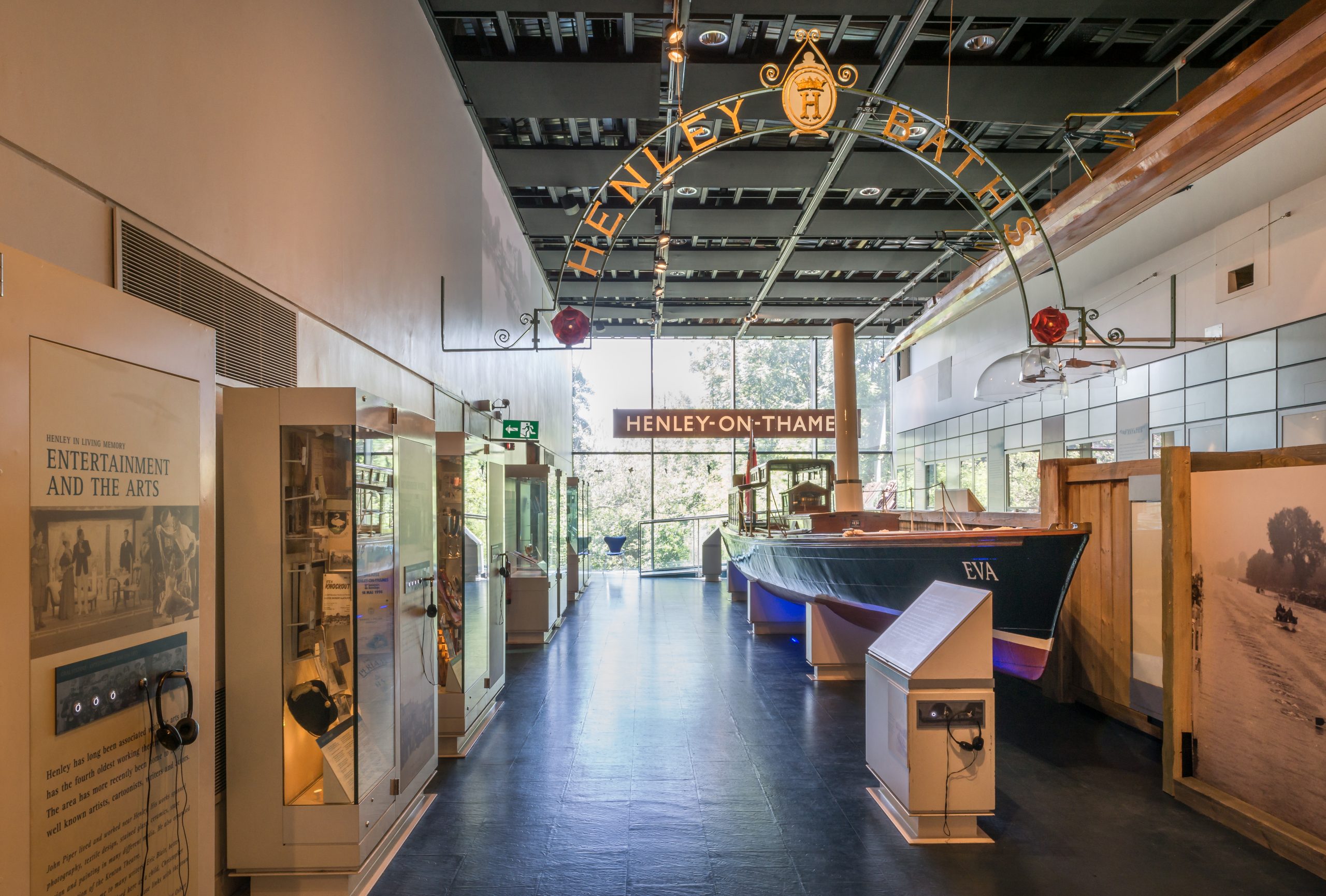 A long view through the Henley Gallery, with display cases on the left wall, a large boat on the right, and large signs for Henley on Thames and Henley Baths at height, before the light window at the end through which trees can be seen.