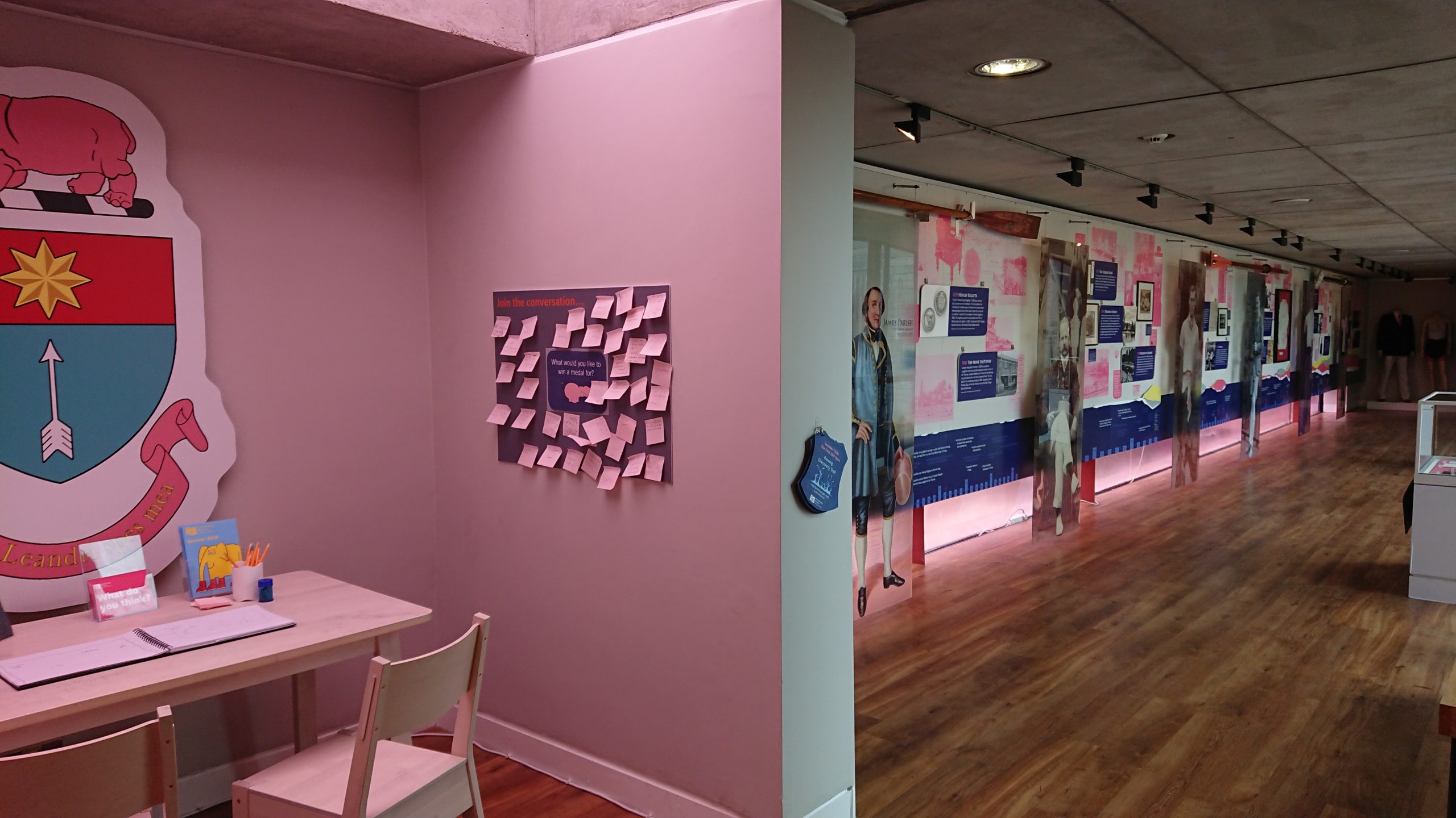 A view down the Community Gallery with a display in pink and navy all about Leander Rowing Club. Life-sized models of rowers are shown alongside trophies in display cases and information on the club. In the foreground is a table and chairs with a comments board.