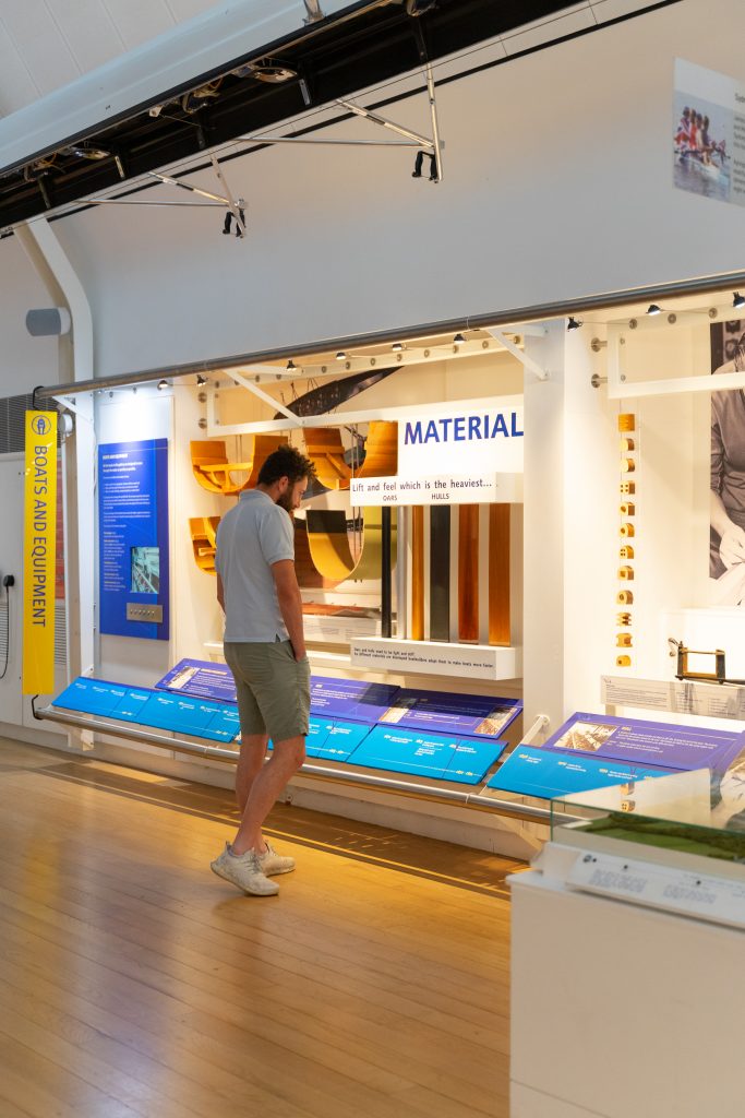 A man wearing shorts stands in front of a display showing the materials that rowing boats and oars are made from. A boat hands above is head.