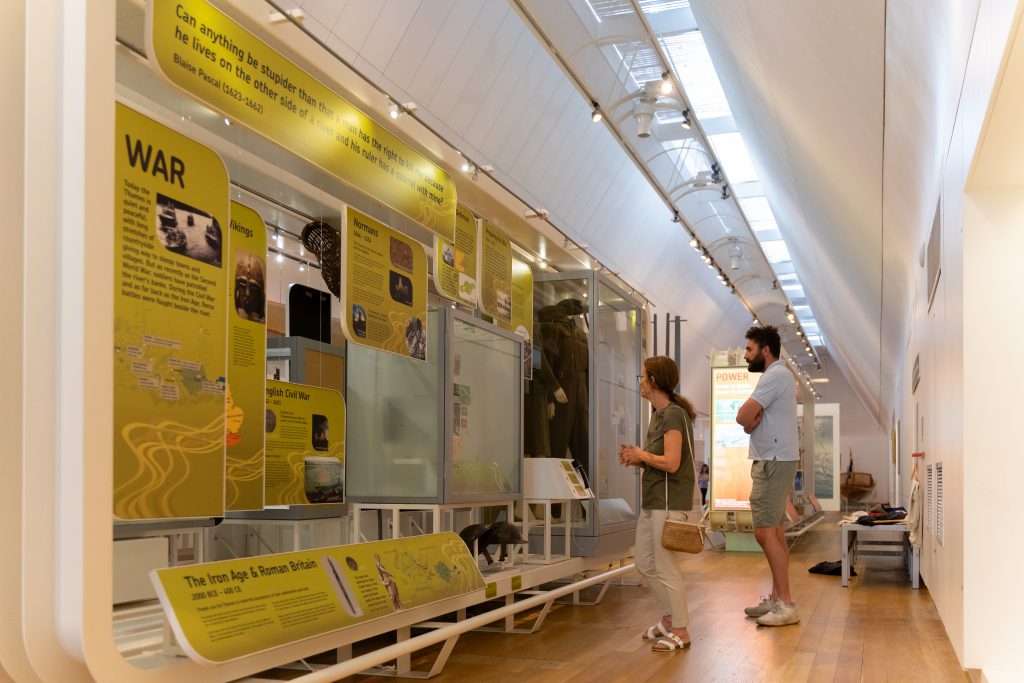 A man and a woman are looking at yellow displays about war in a gallery. There are military uniforms in the case in front of them.