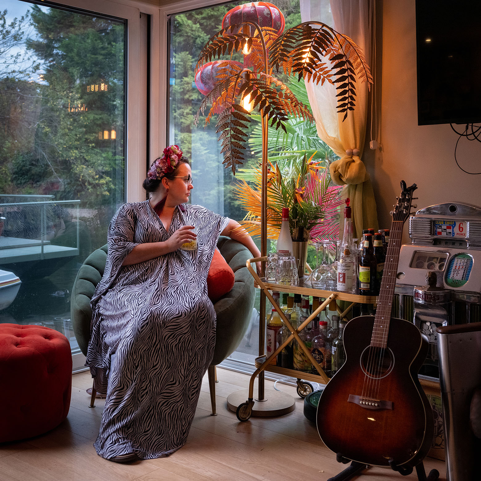 A woman in a zebra print dress sits in a plush chair by a window, holding a glass beneath a palm tree lamp. The room is softly light and there is a drinks cabinet and a guitar to the right.